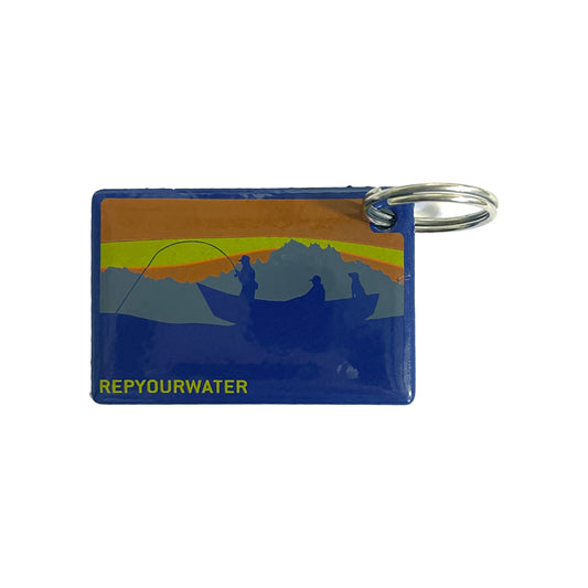 a rectangular key ring shows a scene of people fishing from a drift boat in front of a mountain and sky scene