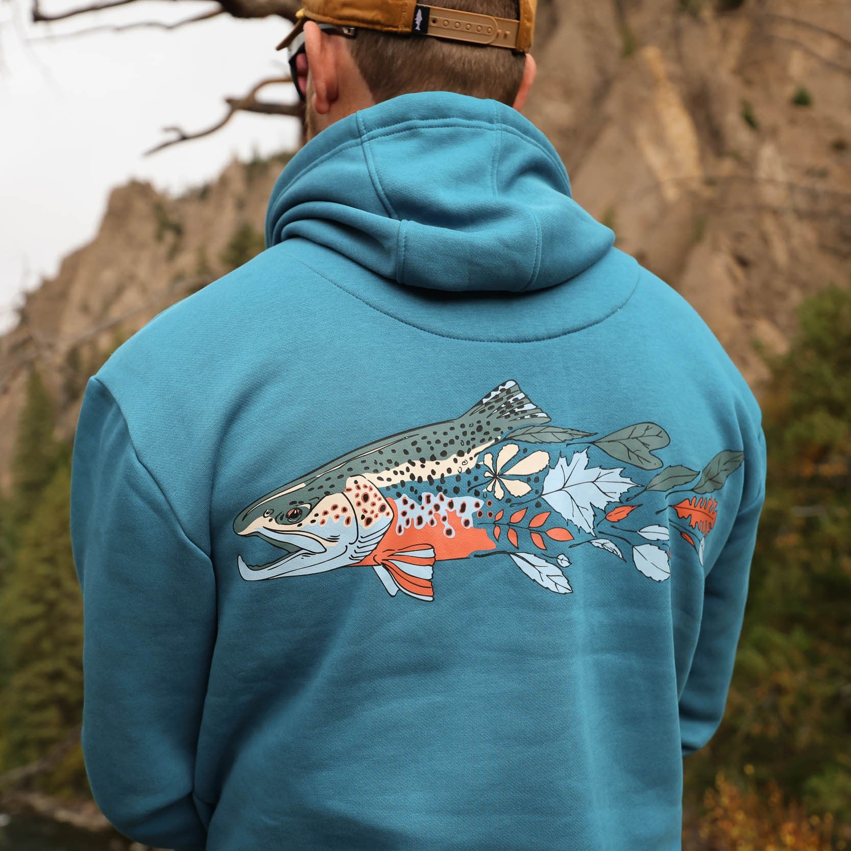 Person wearing a blue hoodie with a fish design in a natural setting. the design is repyourwater fall colors design of a brown trout fading to leaves