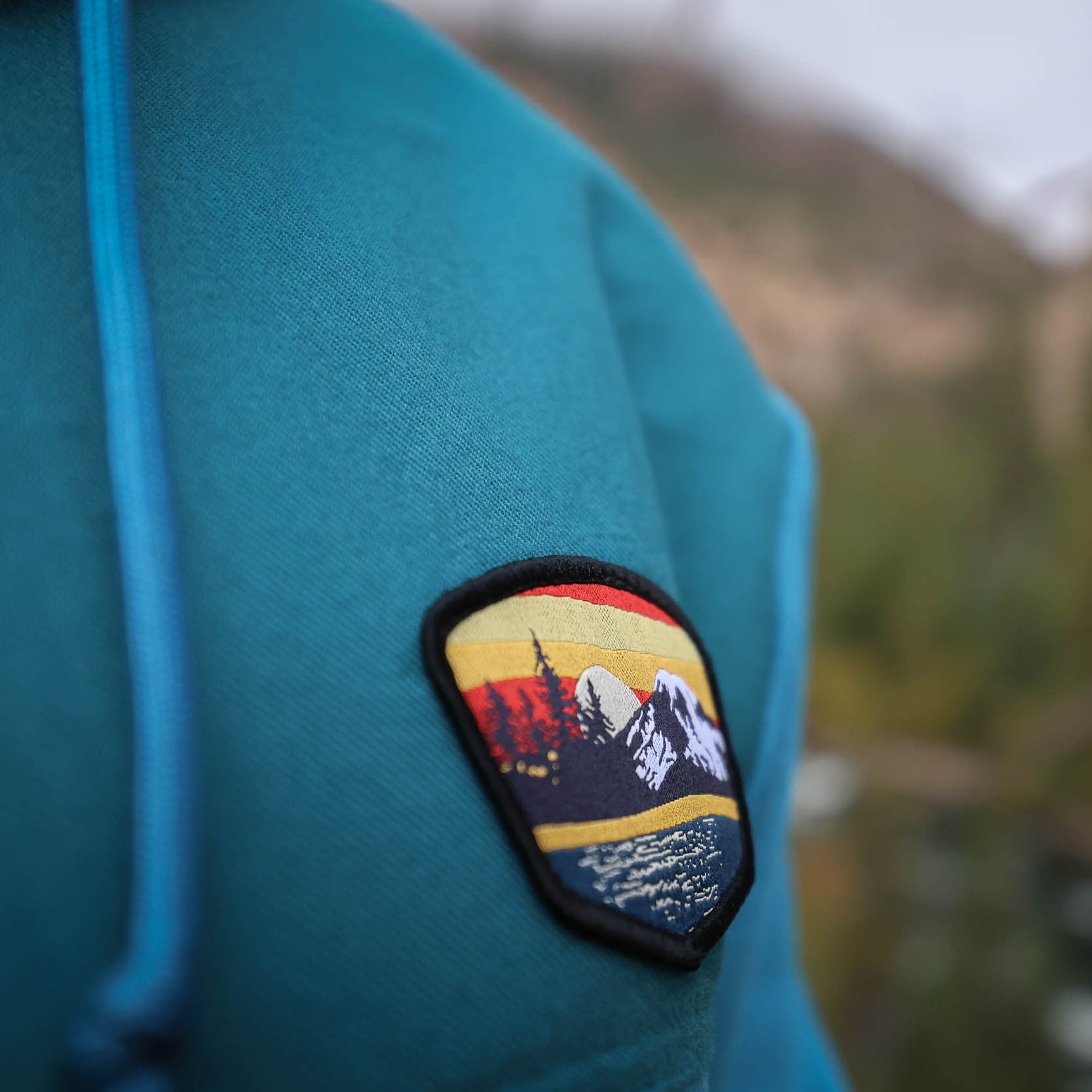 Blue hoody with a mountain sunset-themed patch against a blurred natural background