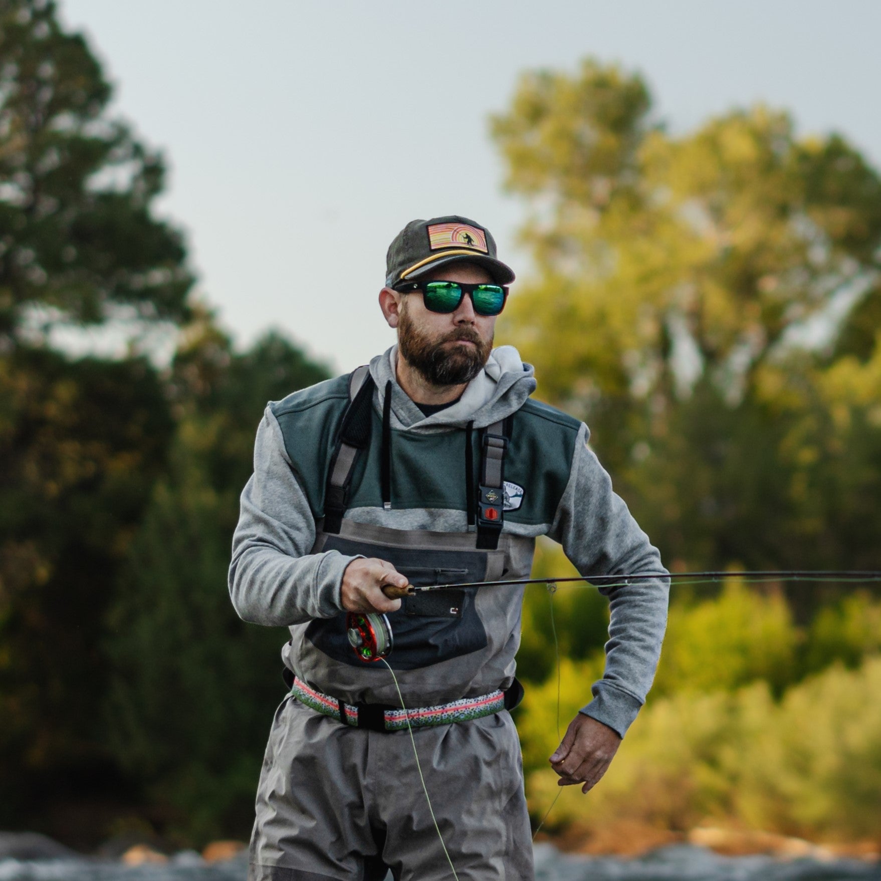 A person fly fishing outdoors, wearing a gray hoodie, fishing vest, chest waders, a baseball cap, and sunglasses, holding a fishing rod with trees and greenery in the background