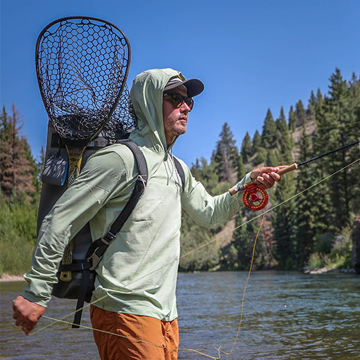 A person fly fishing in a river, wearing a light green hooded shirt, sunglasses, and a backpack with a landing net, holding a fishing rod while standing in shallow water with forested hills in the background.