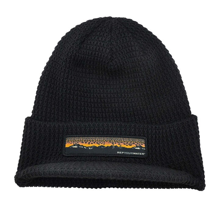 black knit hat with a brim and a patch on the front showing a mountain range and brown trout skin as the "sunset"