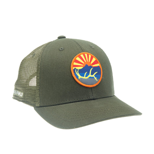 A hat with green mesh in the back with green fabric in the front has a circular patch on front with an elk antler, in front of a mountain and sunset