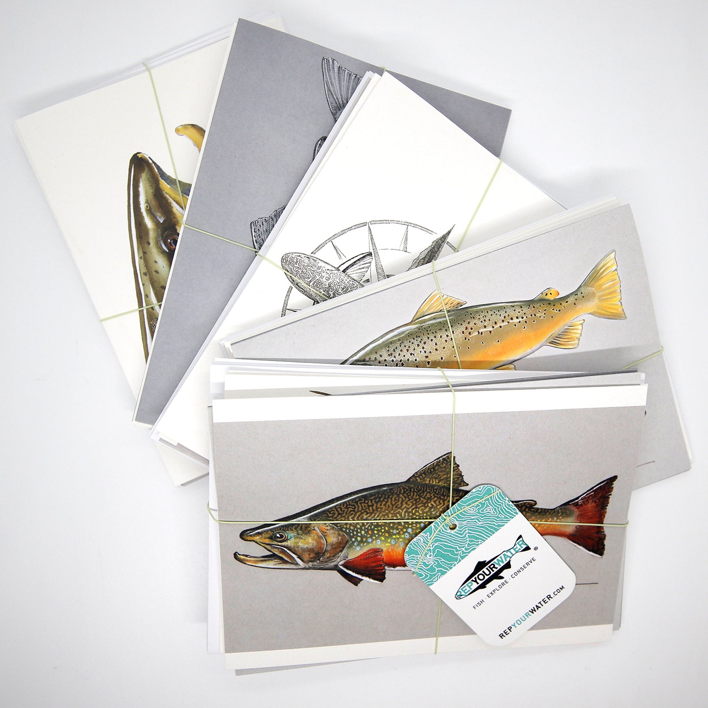 5 stacks of greeting cards in bundles wrapped by fly line and a hangtag on front that reads repyourwater inside a trout silhouette and fish explore conserve. Repyourwater.com is on the hangtag. The top image is a brook trout, then a brown trout, then part of a black and white trout then part of another trout then part of a trout face. All are partially obscured by the one on top of the other