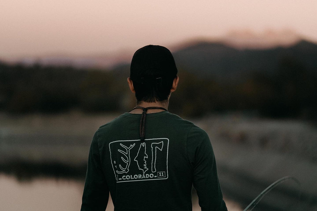 A man looks away from the camera wearing a shirt that reads colorado and has an antler trout and hatchet