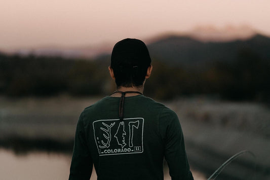 A man looks away from the camera wearing a shirt that reads colorado and has an antler trout and hatchet