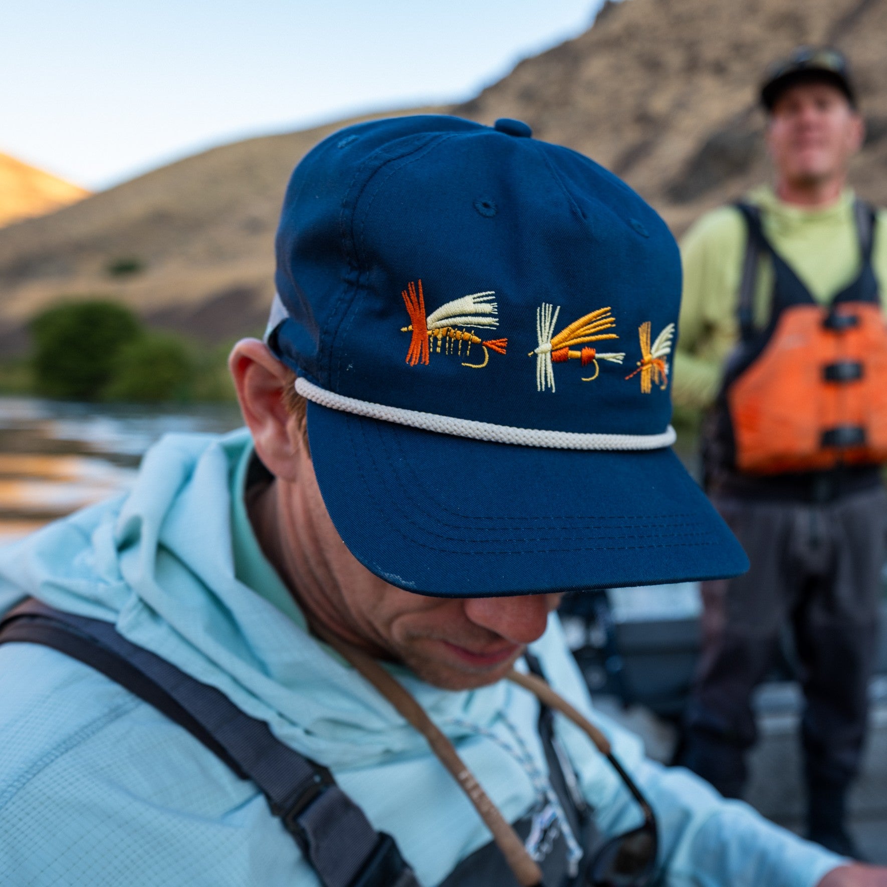 Person wearing a blue cap with fishing fly designs, standing outdoors with mountains in the background.