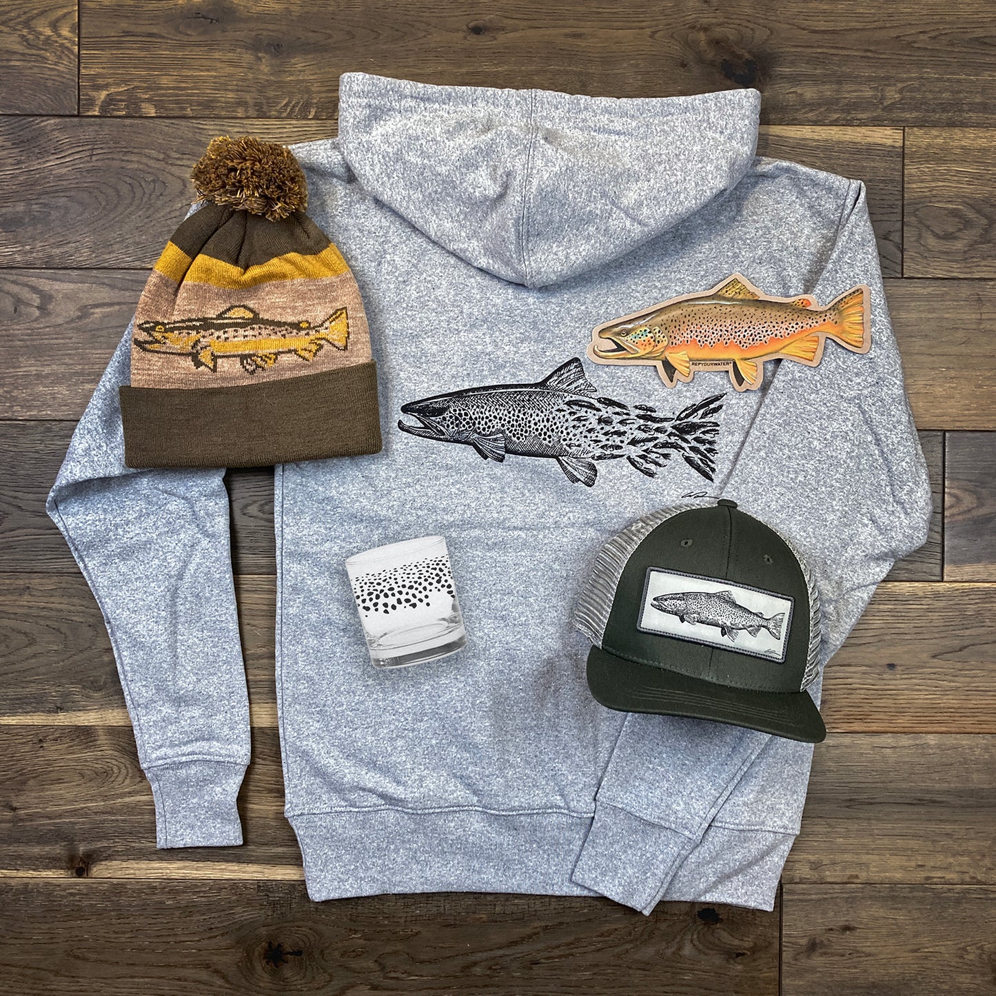 A hooded sweatshirt, knit hat, brimmed hat, glass and sticker A hat, shirt, pair of socks and two stickers laid out on a wood floor.