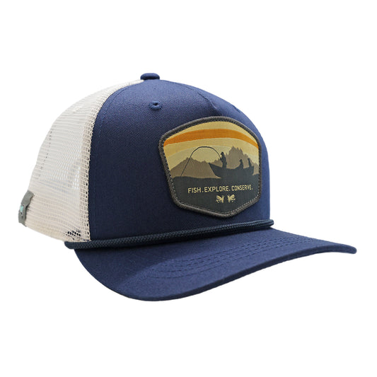 A 5 panel hat with white mesh back and navy blue front. The front design says Fish Explore Conserve and has a boat with anglers and dog, 2 flies, and mountains with sunset skies