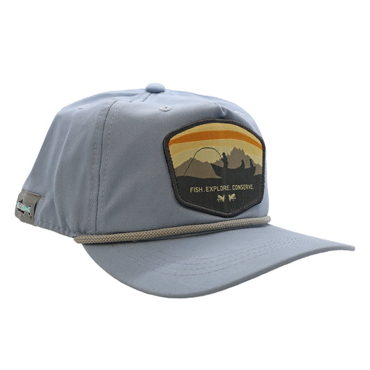 An light blue unstructured hat with a white rope. The front features a design with anglers and dog in a boat, 2 flies, and mountains with sunset skies. It says Fish Explore Conserve.