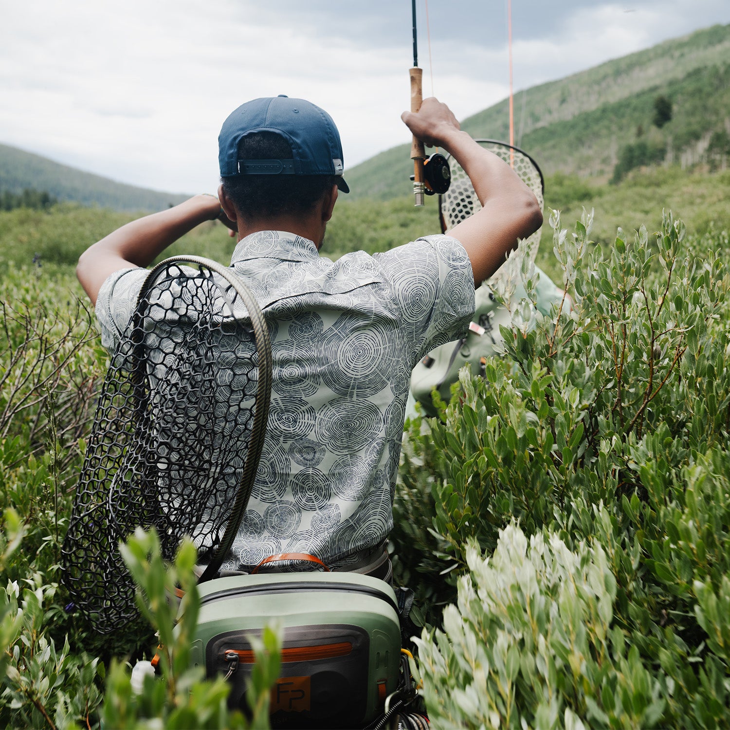 Man fly fishing in a mountain valley, wearing a patterned short sleeve shirt and carrying a net and gear pack