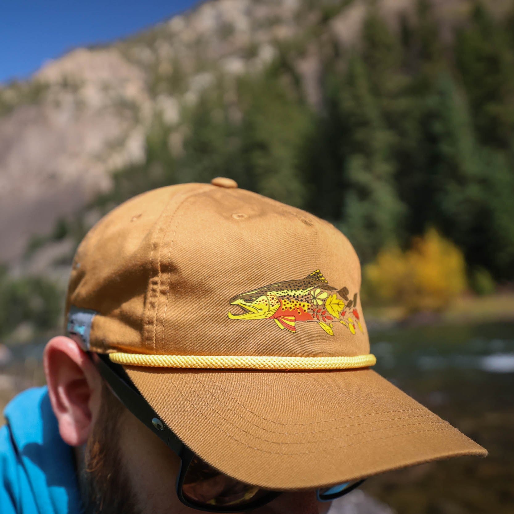 Person wearing a brown hat with the repyourwater fall colors design that shows a brown trout fading to colored leaves near a mountain and river.