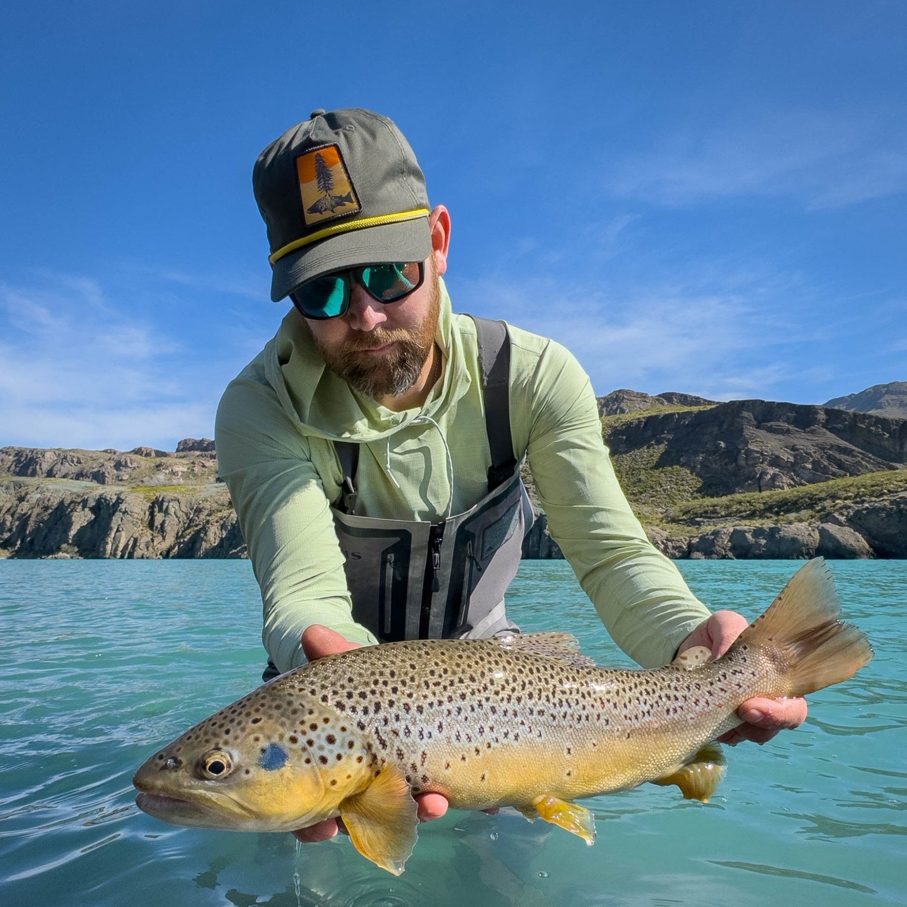 man wearing Green full cloth hat with a patch of a tree and trout silhouette and yellow rope and holding a brown trout in glacial water