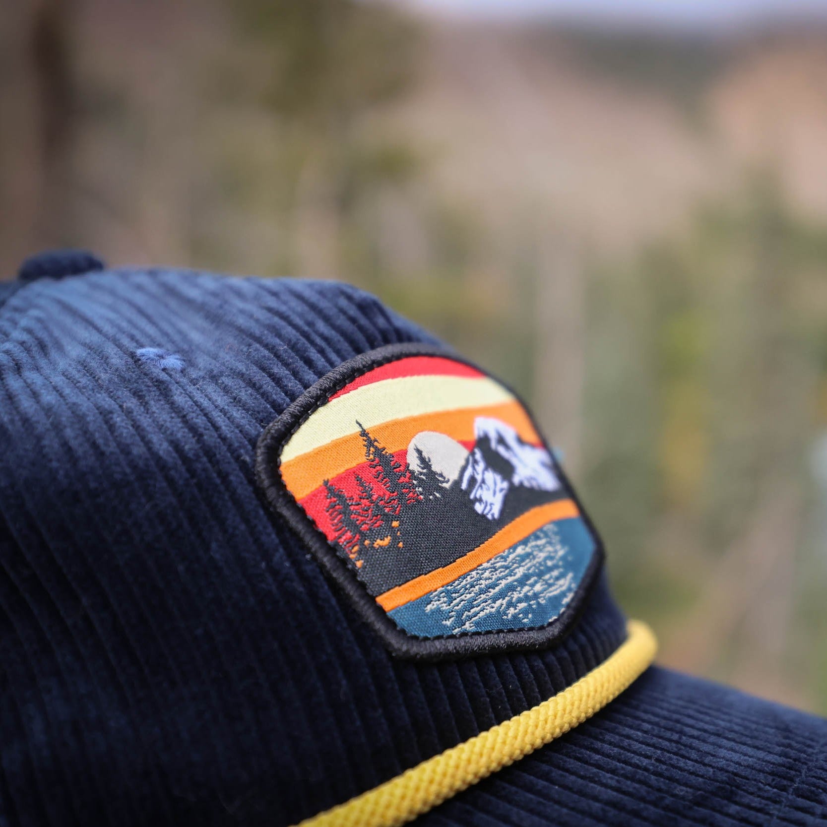 Navy blue corduroy cap with a mountain and tree sunset patch against a blurred natural background