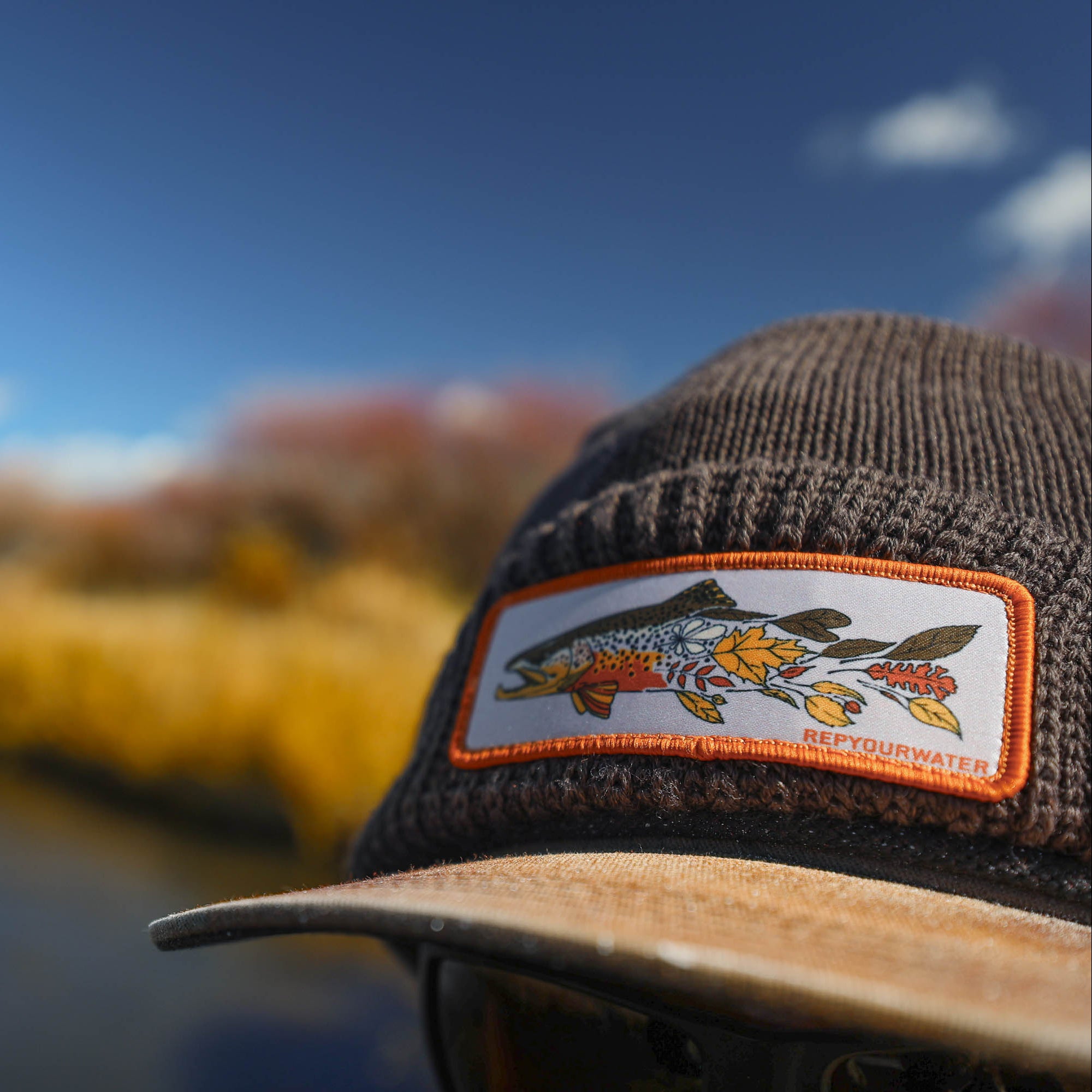 Close-up of a brown knit cap with a stitched patch showing a trout and leaves and the text “RepYourWater,” with a blurred river and autumn foliage in the background.