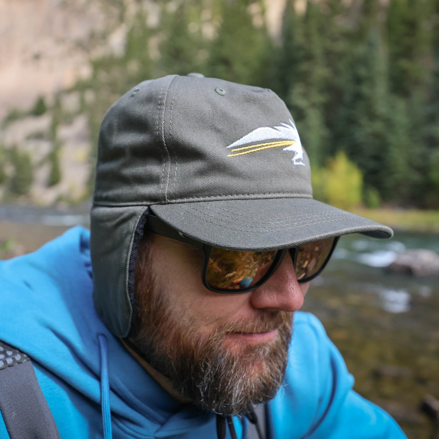 Man wearing a cap and sunglasses outdoors near a body of water the hat is dark green with ear flaps and a streamer fly design on the front