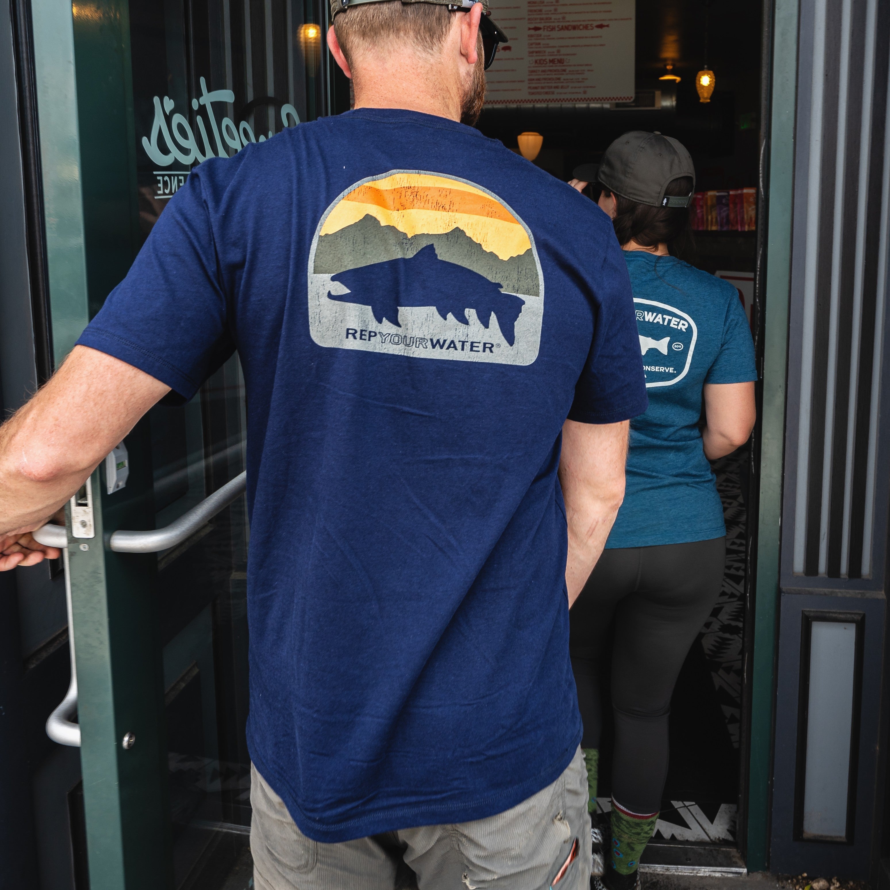 Two people enter a restaurant; the foreground person wears a blue T-shirt with a fish and mountain graphic and the words ‘Rep Your Water,’ holding the door open as the second person walks inside.