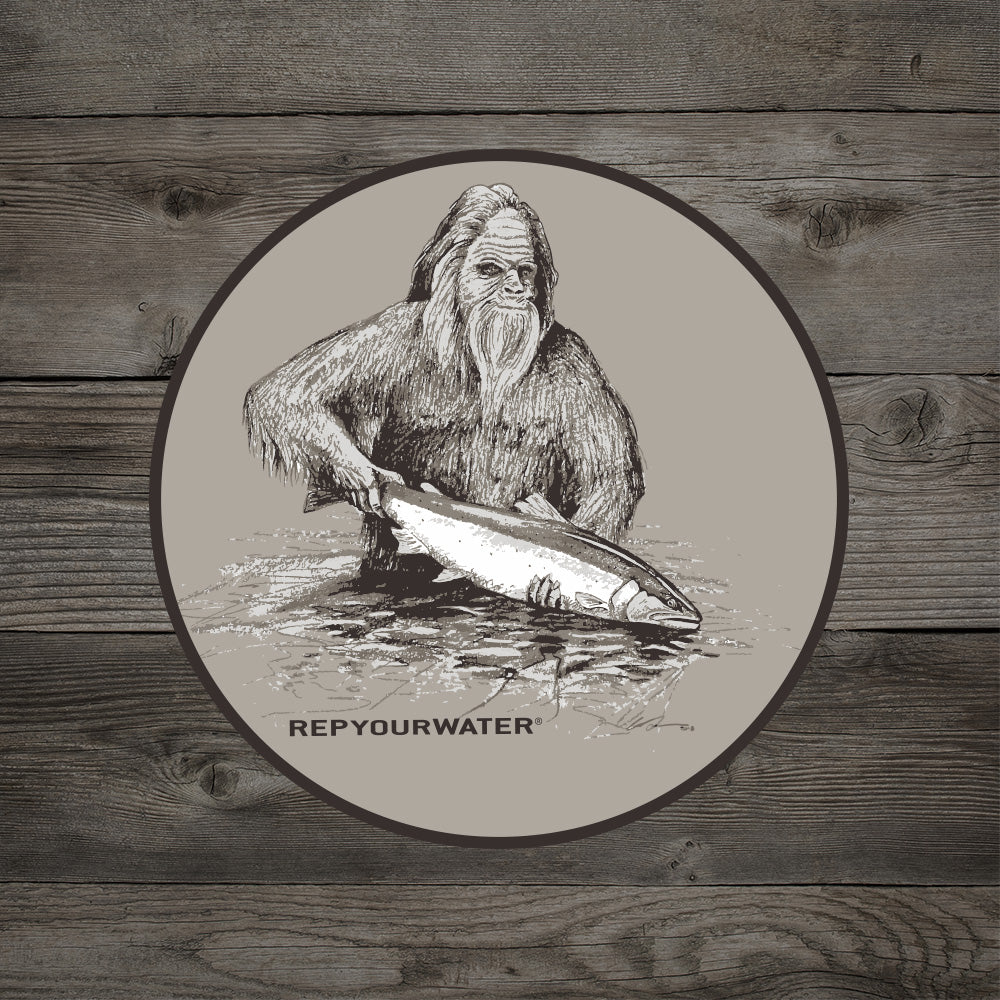 A sticker on a wooden background features drawing of a sasquatch holding a fish in the water