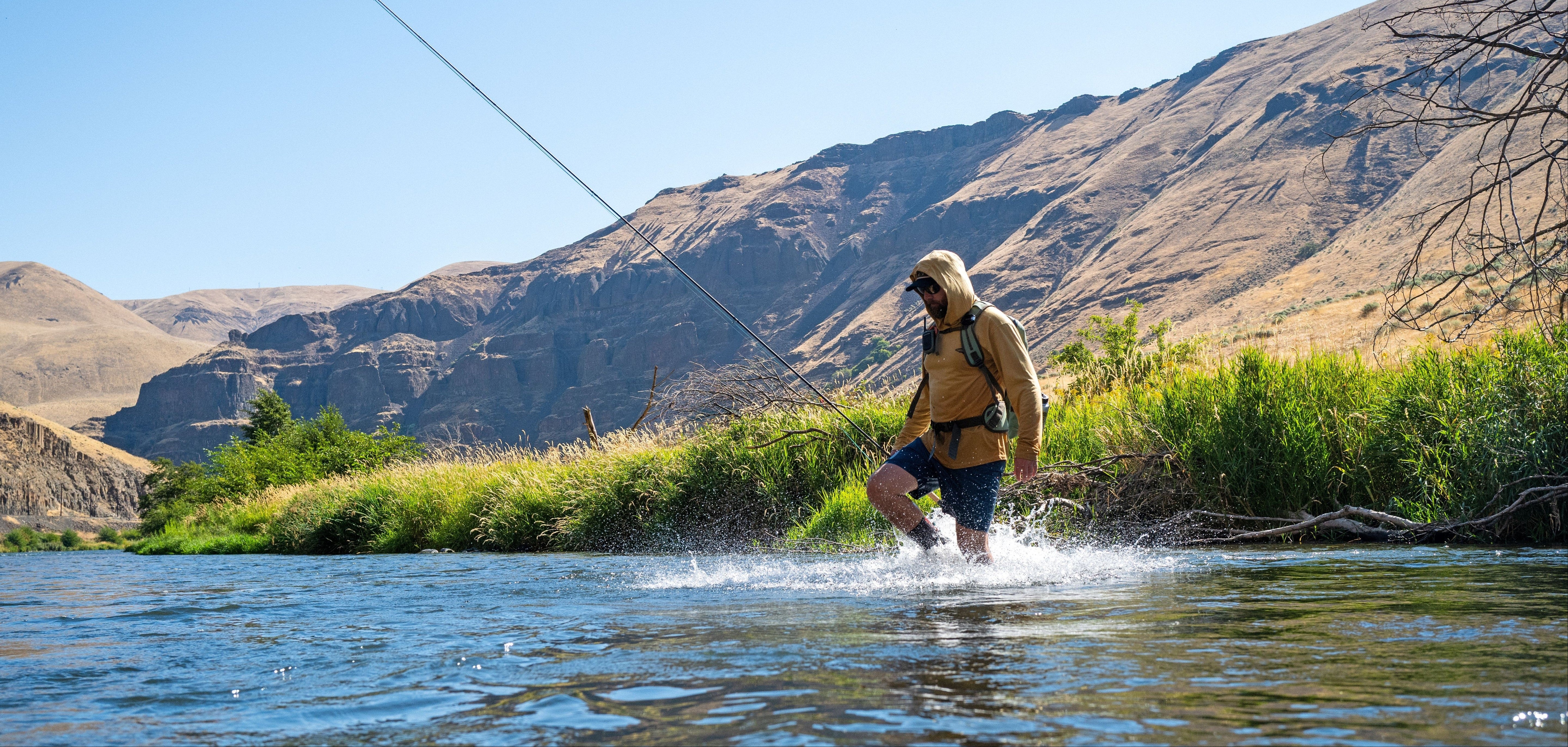 A person wearing a hooded sun shirt and backpack wades through a river while fly fishing, with water splashing around their legs. Dry, rugged mountains rise in the background under a clear blue sky.