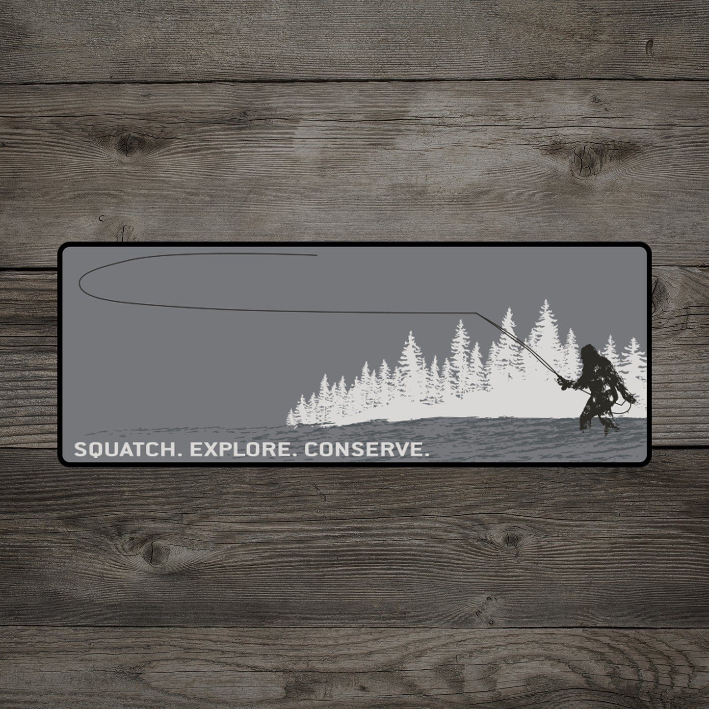 A wood background has a sticker on it that features a sasquatch casting a fly rod while in the water in front of pine trees with the words squatch explore conserve underneath