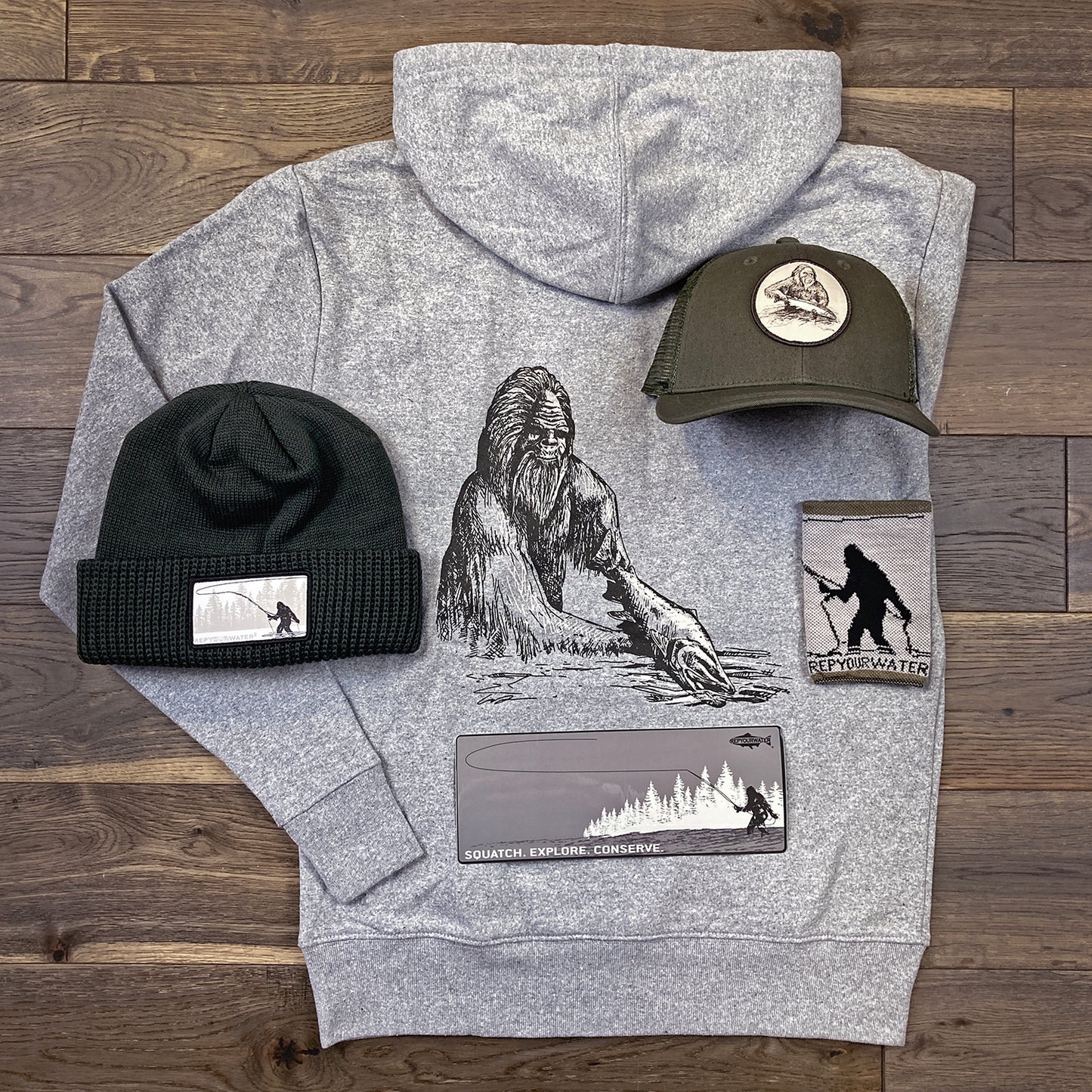 A knit hat, brimmed hat, hooded sweatshirt, can cooler, and sticker laid out on a wood floor.