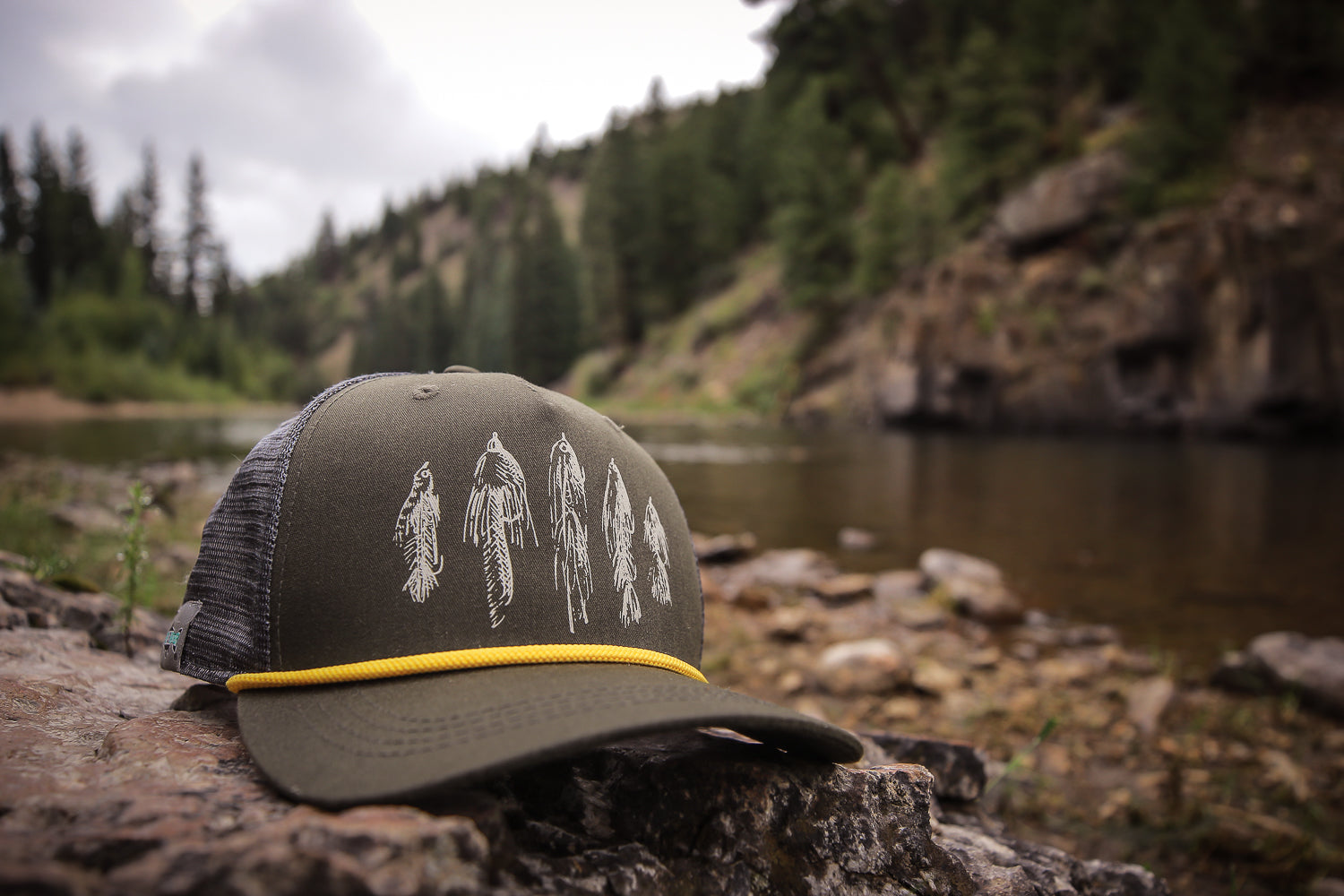 A hat on on a rock by a river - the hat has streamer flies on the front.