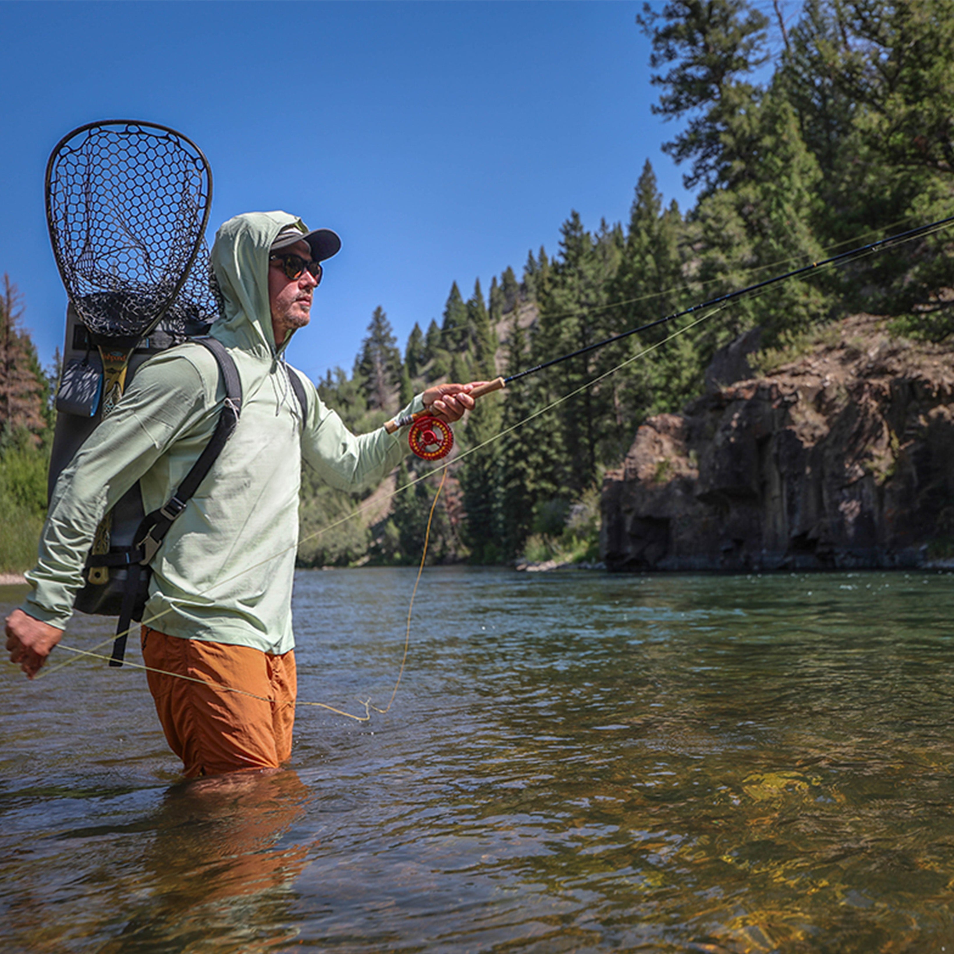 Person fly fishing while wading in a river, wearing a sun hoodie, hat, and backpack with a net, casting toward a rocky, tree-lined shoreline under a clear blue sky.