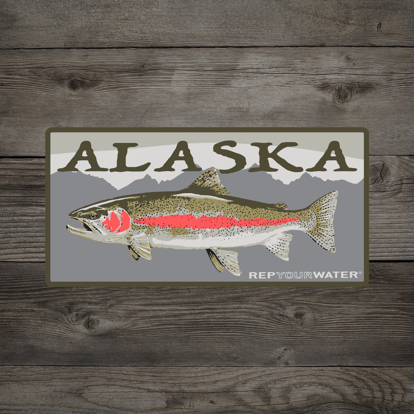 A wooden background with a mockup of a sticker is shown.  The sticker says alaska with a rainbow trout on it