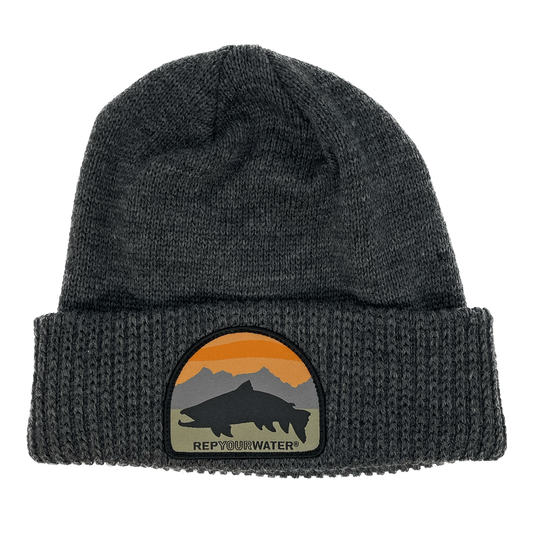 A gray winter hat with a cuff and a patch on the cuff with a mountain range background with a black trout silhouette