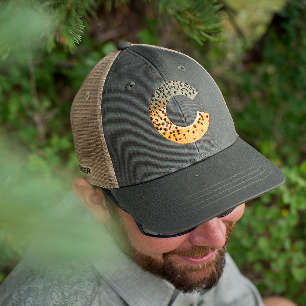 A man smiles and wears the Colorado brown trout skin hat with trees behind him