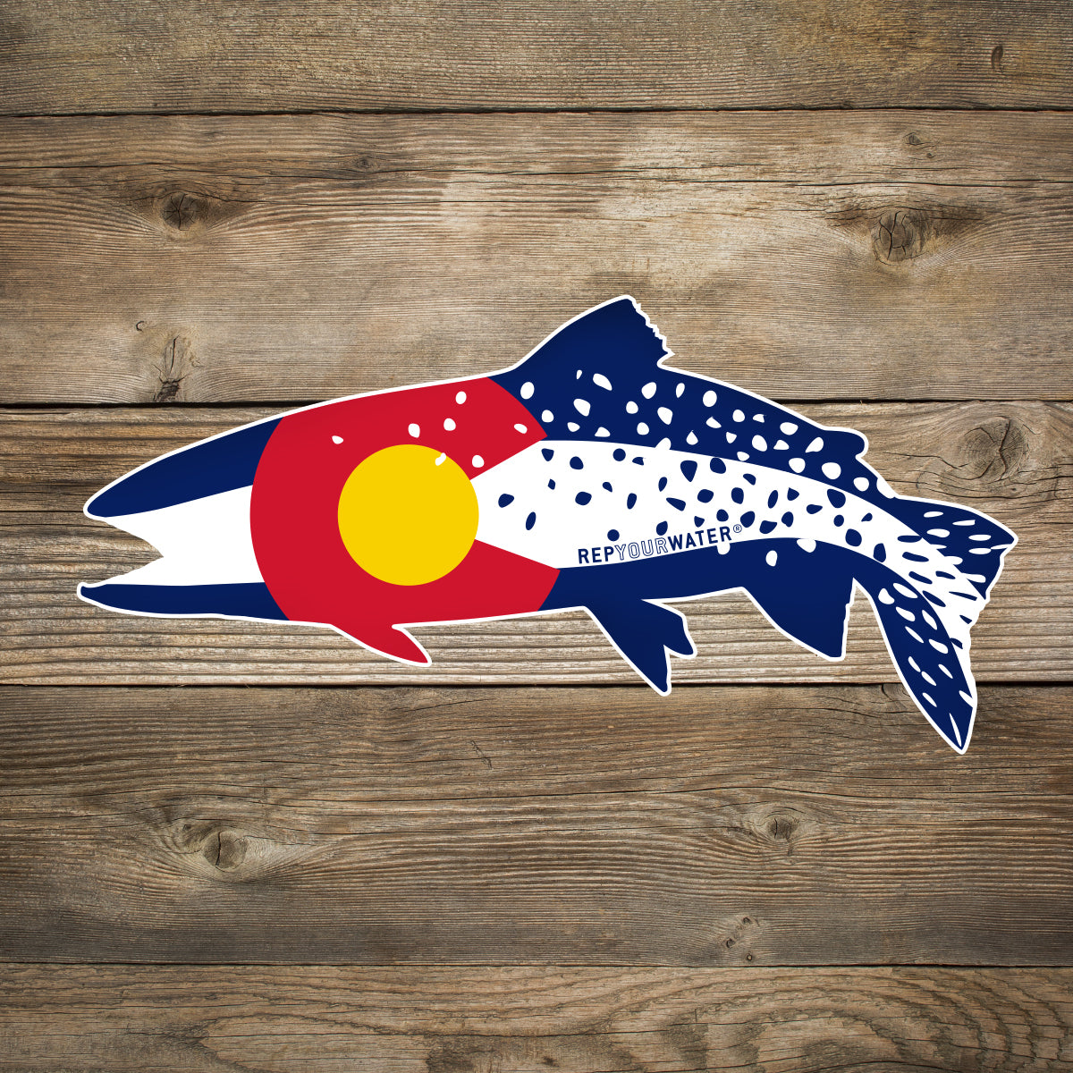 A wood background with a mockup of a sticker in the shape of a trout and the colors and pattern of the colorado flag