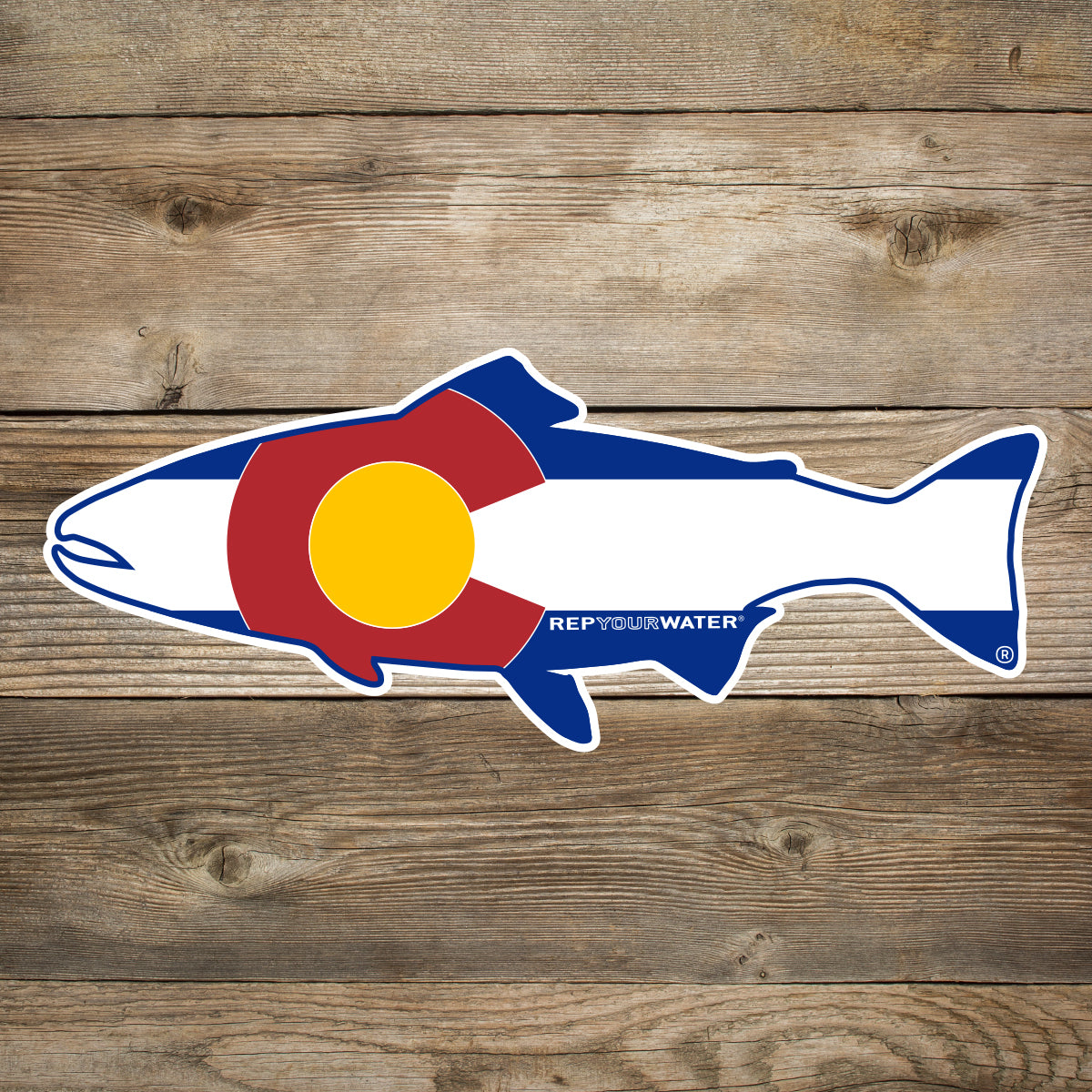 A wood background with a mockup of a sticker in the shape of a trout with the colors and pattern of the colorado flag