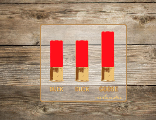 A wooden background shows the mockup of a sticker that shows 3 shot gun shells that says duck duck goose below them