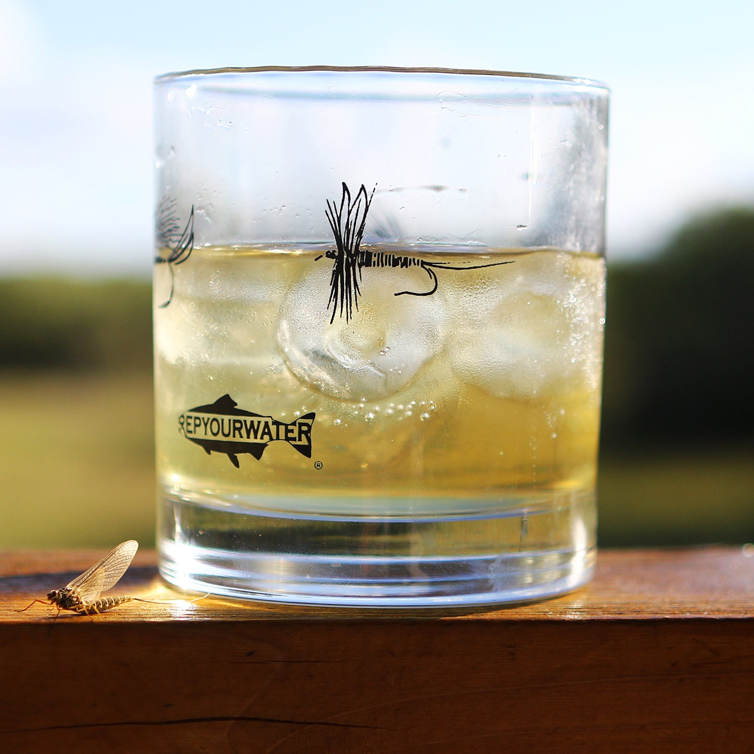A low ball cocktail glass is on a wood railing.  The glass has brown liquid and ice in it and has a printed dry fly on it with a logo that reads repyourwater inside a trout silhouette. A real mayfly is next to the glass