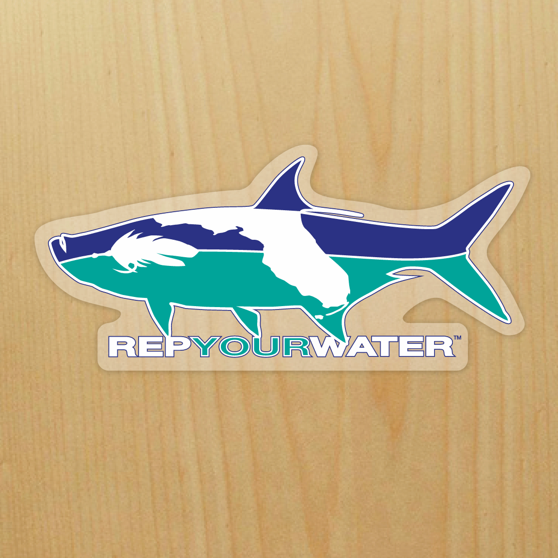 A sticker is shown on a wood background - the sticker is a tarpon shape in blue and turquoise with a fly and the state shape of Florida inside of the fish.