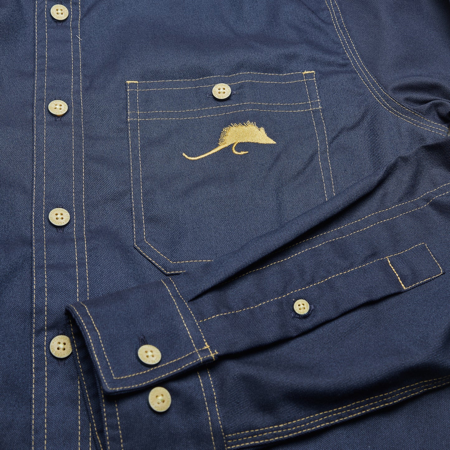 Detail photo of the front pocket and cuff/sleeve of a navy blue shirt with a fly embroidered on the pocket.