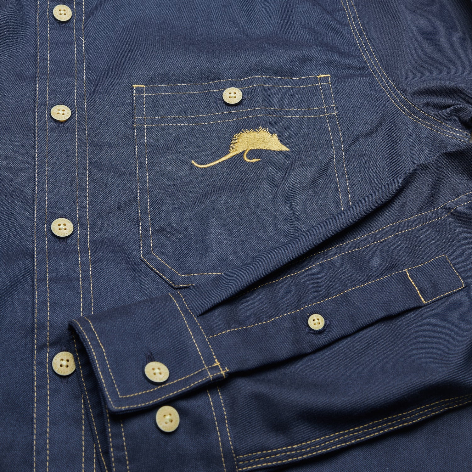 Detail photo of the front pocket and cuff/sleeve of a navy blue shirt with a fly embroidered on the pocket.