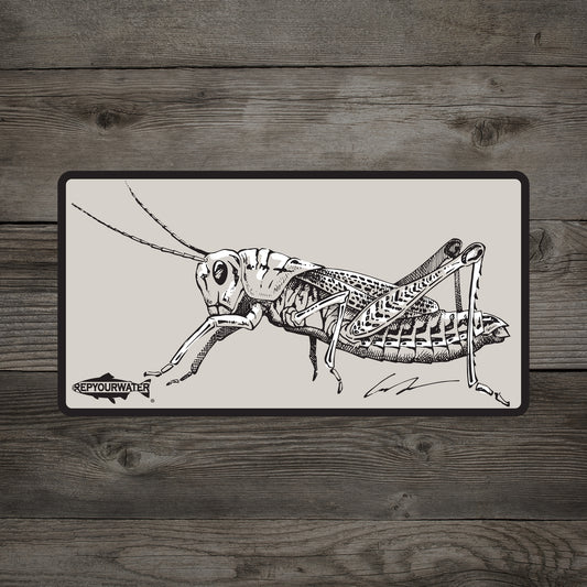 A rectangular sticker on a wood background. The sticker features a drawing of a grasshopper on it.