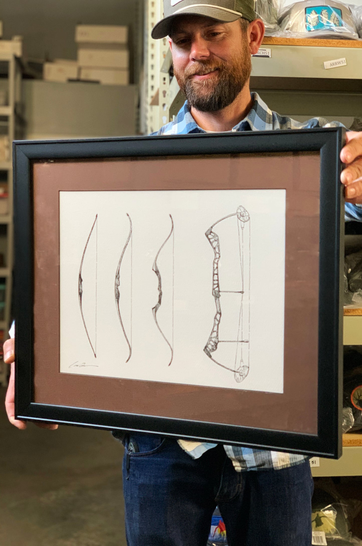 A man holds a framed piece of art which is a drawing of four hunting bows. Three are traditional long bows and the last one is a modern compound bow