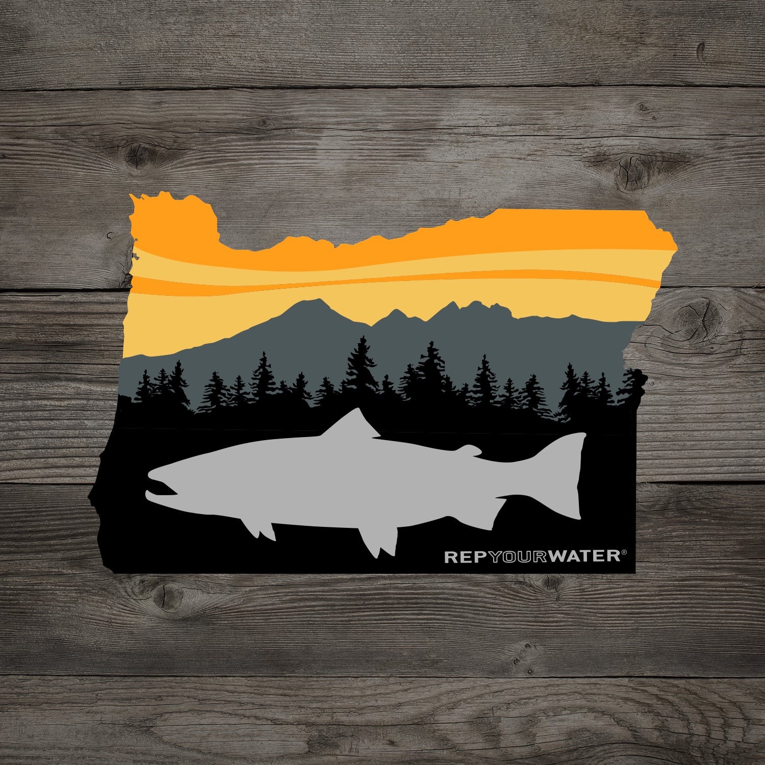 A wood background has a sticker in the shape of oregon stsate with a trout in front of trees mountains and a sunset