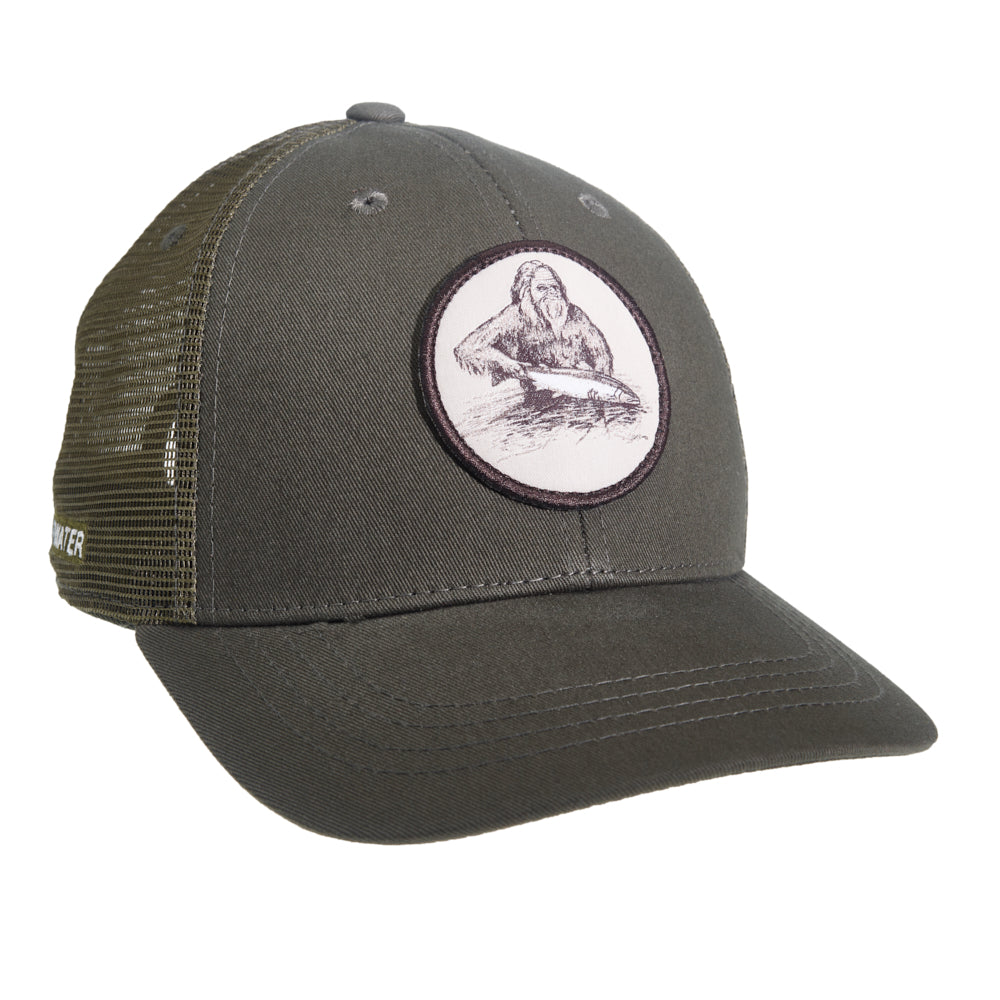 A hat with green mesh in back and green fabric in front features a circular patch which shows drawing of a sasquatch holding a fish in the water