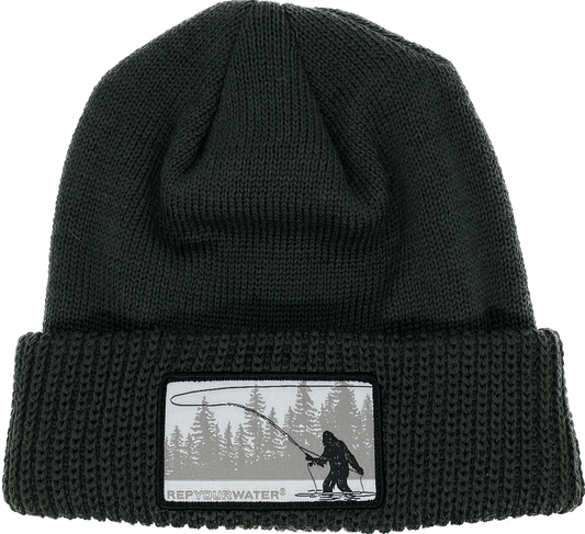 A dark forest green winter hat with a cuff and a rectangular patch of a sasquatch casting with a line of trees in the background