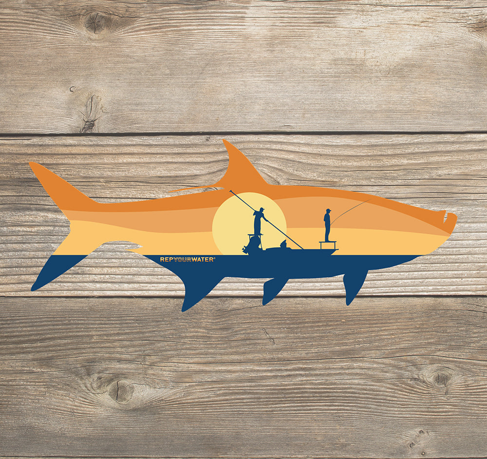 A sticker on a wood background that features tarpon shape with a boat and sunrise in side it