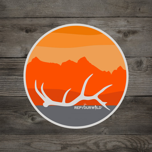 A wooden background has a sticker on it featuring an antler in front of a mountain and sunset scene and reads repyourwild
