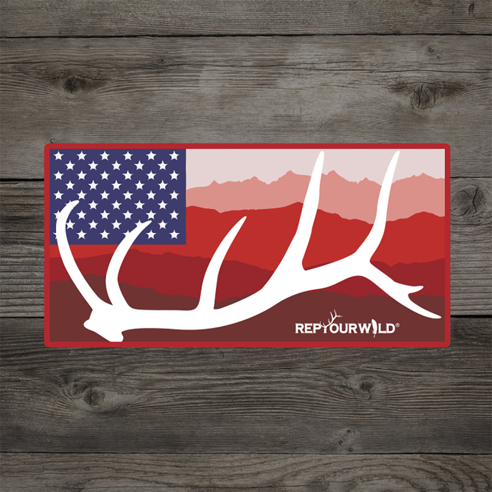 A sticker with an elk antler in front of mountains and 50 stars in a blue rectangle on a wood background